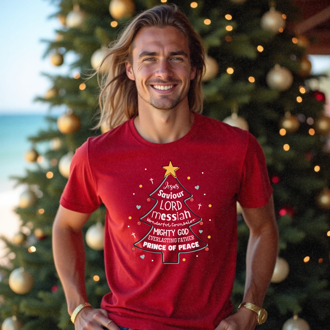 Young male with long blonde hair wearing a red Christmas t-shirt in front of a Christmas tree near the beach with 'Jesus Saviour Lord Messiah' on the front by Faith Looks Good