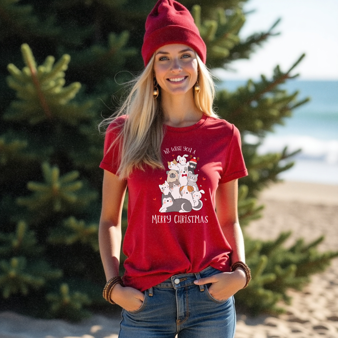 Young girl in a Christmas beanie standing on the beach in front of a Christmas tree with long blonde hair wearing a funny red Christmas t-shirt with a pile of cats and dogs in the shape of a Christmas tree and words 'We Wish You A Merry Christmas' surrounding the graphic by Faith Looks Good