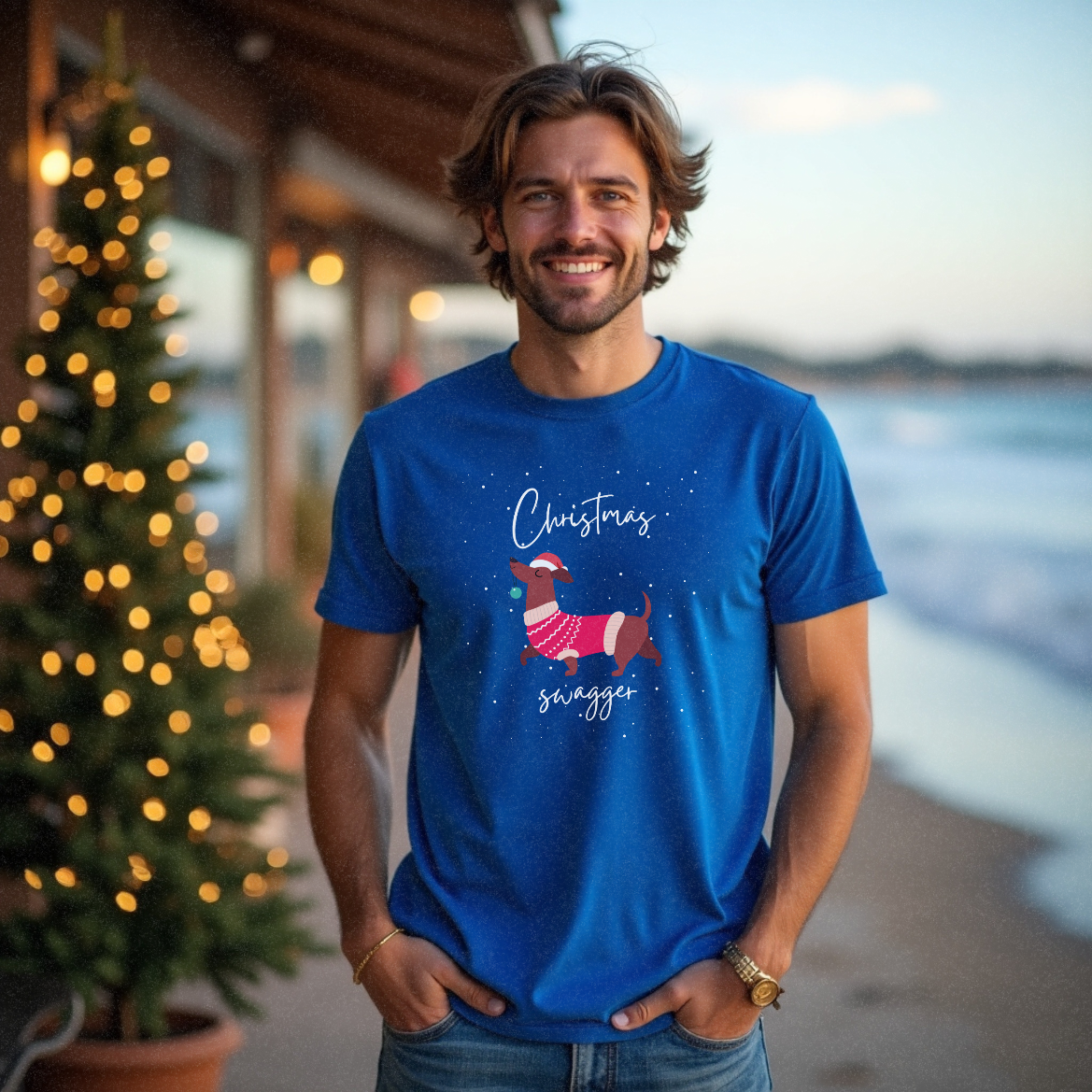 Young male in his 20s with short hair standing near a beach restaurant wearing a funny blue Christmas t-shirt with a graphic of a daschund wearing a beaning and carrying a bauble in its mouth and nose in the air with the text 'Christmas Swagger' by Faith Looks Good
